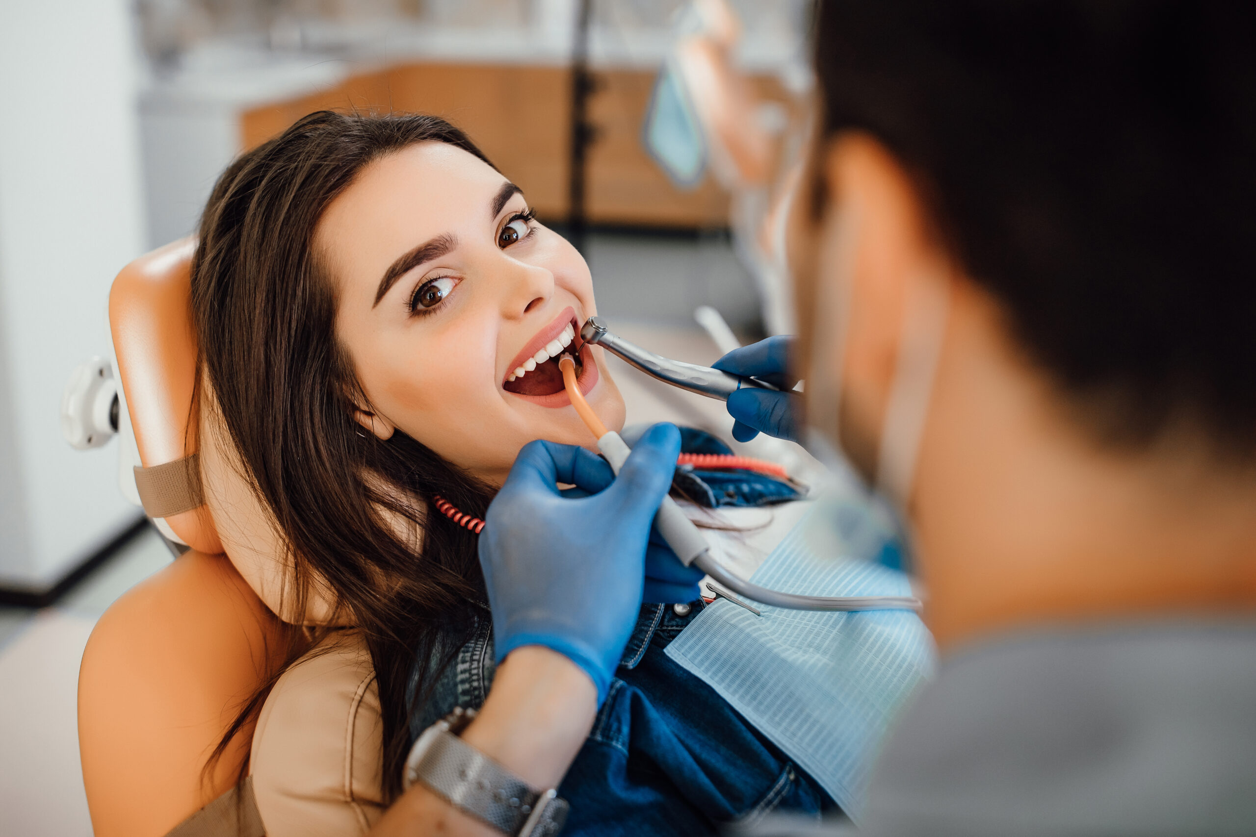 young-female-patient-visiting-dentist-office Young female patient visiting dentist office. Beautiful woman with healthy straight white teeth sitting at dental chair with open mouth during oral checkup while doctor working at teeth.